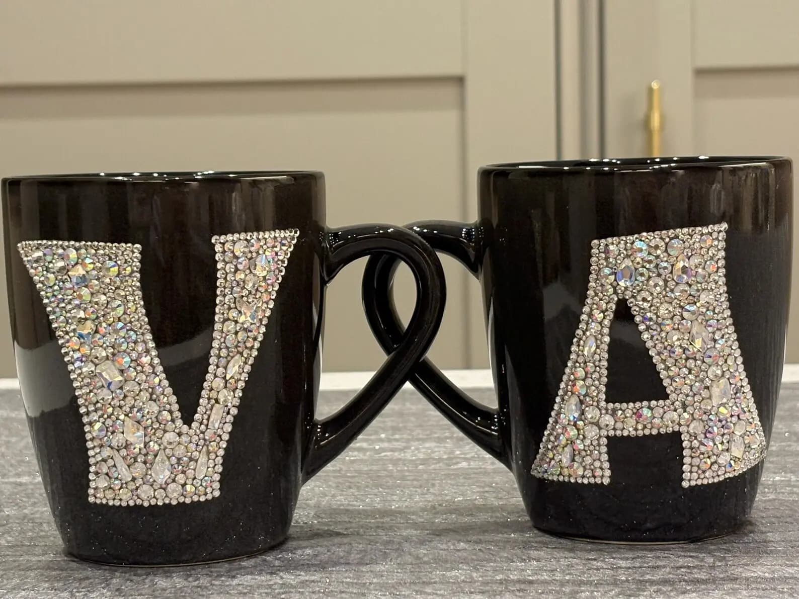 Personalized Crystal Initial Mug with letters "V" and "A" in sparkling crystals, ceramic, 450ml, handles intertwined, on a table.