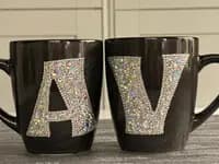 Personalized Crystal Initial Mug, black ceramic with "A" and "V" in sparkling crystals, displayed side by side on a textured surface.