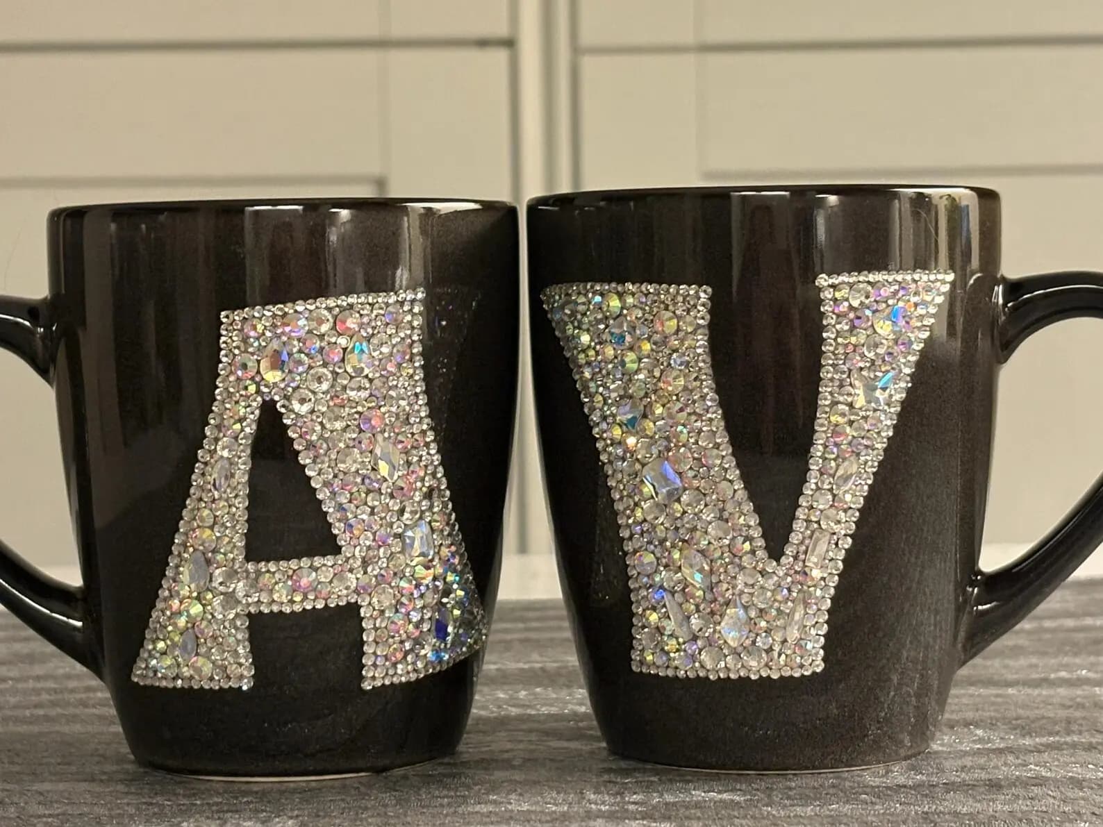 Personalized Crystal Initial Mug, black ceramic with "A" and "V" in sparkling crystals, displayed side by side on a textured surface.